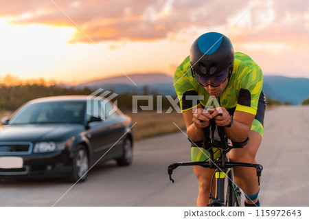 Close up photo of triathlete riding his bicycle during sunset, preparing for a marathon. The warm colors of the sky provide a beautiful backdrop for his determined and focused effort. Close up photo of triathlete riding his bicycle during sunset, preparing for a marathon. The warm colors of the sky provide a beautiful backdrop for his determined and focused effort. 115972643