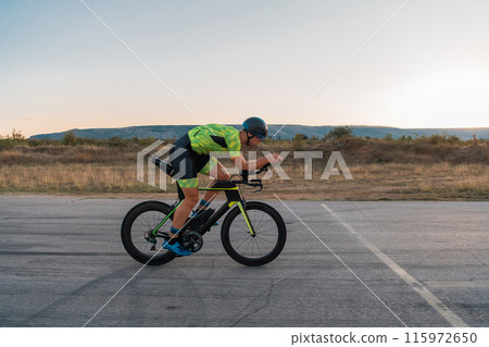 Triathlete riding his bicycle during sunset, preparing for a marathon. The warm colors of the sky provide a beautiful backdrop for his determined and focused effort. Triathlete riding his bicycle during sunset, preparing for a marathon. The warm colors of the sky provide a beautiful backdrop for his determined and focused effort. 115972650