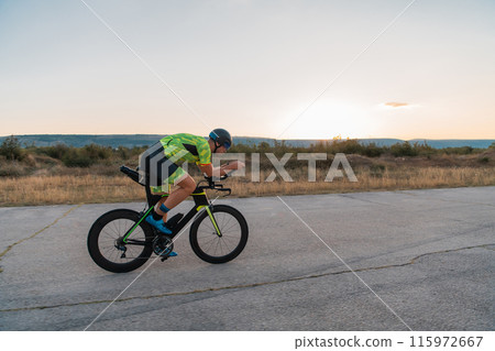 Triathlete riding his bicycle during sunset, preparing for a marathon. The warm colors of the sky provide a beautiful backdrop for his determined and focused effort. 115972667