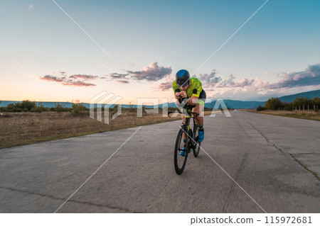 Triathlete riding his bicycle during sunset, preparing for a marathon. The warm colors of the sky provide a beautiful backdrop for his determined and focused effort. 115972681