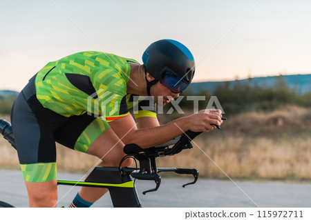 Close up photo of triathlete riding his bicycle during sunset, preparing for a marathon. The warm colors of the sky provide a beautiful backdrop for his determined and focused effort. Close up photo of triathlete riding his bicycle during sunset, preparing for a marathon. The warm colors of the sky provide a beautiful backdrop for his determined and focused effort. 115972711