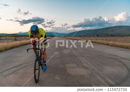 Triathlete riding his bicycle during sunset, preparing for a marathon. The warm colors of the sky provide a beautiful backdrop for his determined and focused effort. 115972730
