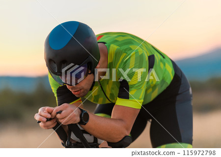 Close up photo of triathlete riding his bicycle during sunset, preparing for a marathon. The warm colors of the sky provide a beautiful backdrop for his determined and focused effort. 115972798