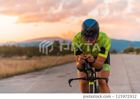 Close up photo of triathlete riding his bicycle during sunset, preparing for a marathon. The warm colors of the sky provide a beautiful backdrop for his determined and focused effort. 115972912