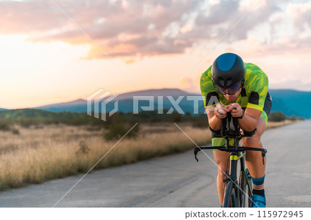 Triathlete riding his bicycle during sunset, preparing for a marathon. The warm colors of the sky provide a beautiful backdrop for his determined and focused effort. 115972945