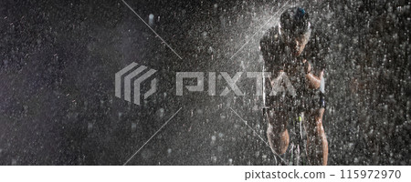 A triathlete braving the rain as he cycles through the night, preparing himself for the upcoming marathon. The blurred raindrops in the foreground and the dark, moody atmosphere in the background add 115972970