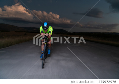 A triathlete rides his bike in the darkness of night, pushing himself to prepare for a marathon. The contrast between the darkness and the light of his bike creates a sense of drama and highlights the A triathlete rides his bike in the darkness of night, pushing himself to prepare for a marathon. The contrast between the darkness and the light of his bike creates a sense of drama and highlights the 115972976