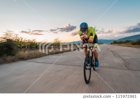 Triathlete riding his bicycle during sunset, preparing for a marathon. The warm colors of the sky provide a beautiful backdrop for his determined and focused effort. Triathlete riding his bicycle during sunset, preparing for a marathon. The warm colors of the sky provide a beautiful backdrop for his determined and focused effort. 115972981