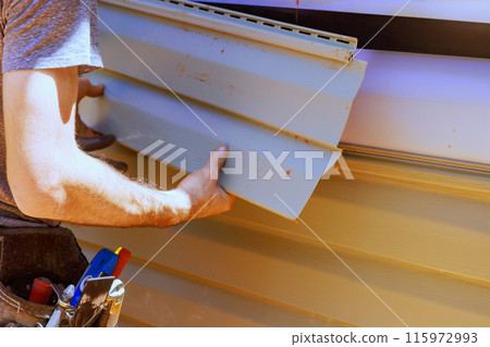 As part of construction, worker installs vinyl plastic siding panels on house facade 115972993