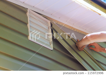 During construction, an employee installs vinyl plastic siding panels on house facade 115972996