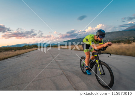 Triathlete riding his bicycle during sunset, preparing for a marathon. The warm colors of the sky provide a beautiful backdrop for his determined and focused effort. Triathlete riding his bicycle during sunset, preparing for a marathon. The warm colors of the sky provide a beautiful backdrop for his determined and focused effort. 115973010