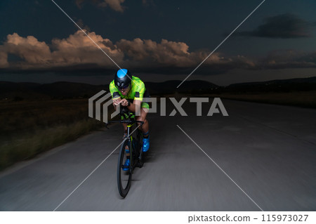 A triathlete rides his bike in the darkness of night, pushing himself to prepare for a marathon. The contrast between the darkness and the light of his bike creates a sense of drama and highlights the 115973027