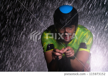 A triathlete braving the rain as he cycles through the night, preparing himself for the upcoming marathon. The blurred raindrops in the foreground and the dark, moody atmosphere in the background add 115973152