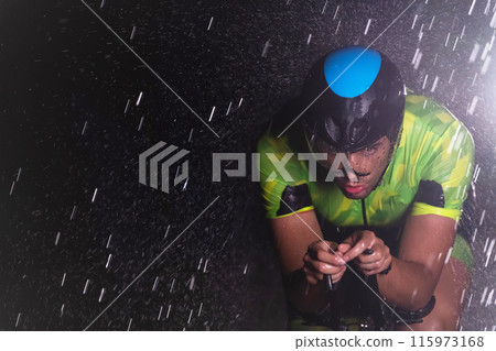 A triathlete braving the rain as he cycles through the night, preparing himself for the upcoming marathon. The blurred raindrops in the foreground and the dark, moody atmosphere in the background add 115973168