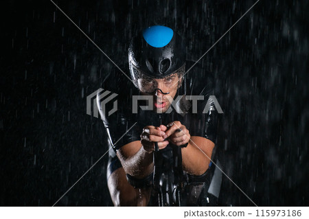 A triathlete braving the rain as he cycles through the night, preparing himself for the upcoming marathon. The blurred raindrops in the foreground and the dark, moody atmosphere in the background add A triathlete braving the rain as he cycles through the night, preparing himself for the upcoming marathon. The blurred raindrops in the foreground and the dark, moody atmosphere in the background add 115973186