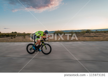 Triathlete riding his bicycle during sunset, preparing for a marathon. The warm colors of the sky provide a beautiful backdrop for his determined and focused effort. 115973191