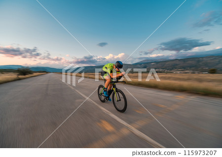 Triathlete riding his bicycle during sunset, preparing for a marathon. The warm colors of the sky provide a beautiful backdrop for his determined and focused effort. 115973207