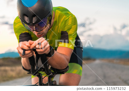Close up photo of triathlete riding his bicycle during sunset, preparing for a marathon. The warm colors of the sky provide a beautiful backdrop for his determined and focused effort. 115973232