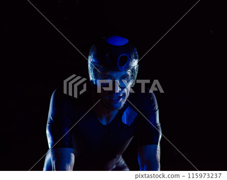 A triathlete rides his bike in the darkness of night, pushing himself to prepare for a marathon. The contrast between the darkness and the light of his bike creates a sense of drama and highlights the A triathlete rides his bike in the darkness of night, pushing himself to prepare for a marathon. The contrast between the darkness and the light of his bike creates a sense of drama and highlights the 115973237