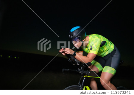 A triathlete rides his bike in the darkness of night, pushing himself to prepare for a marathon. The contrast between the darkness and the light of his bike creates a sense of drama and highlights the A triathlete rides his bike in the darkness of night, pushing himself to prepare for a marathon. The contrast between the darkness and the light of his bike creates a sense of drama and highlights the 115973241