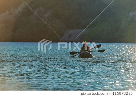 A young couple enjoying an idyllic kayak ride in the middle of a beautiful river surrounded by forest greenery in sunset time  115973255
