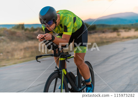 Close up photo of triathlete riding his bicycle during sunset, preparing for a marathon. The warm colors of the sky provide a beautiful backdrop for his determined and focused effort. 115973282