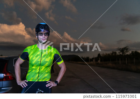 A triathlete resting on the road after a tough bike ride in the dark night, leaning on his bike in complete exhaustion  115973301