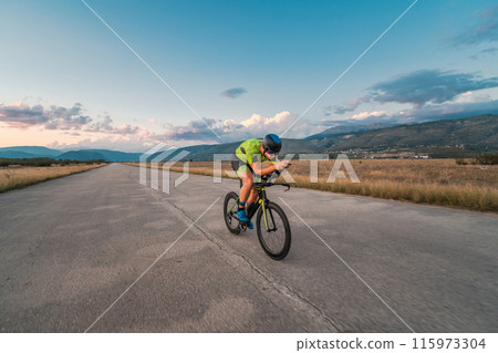 Triathlete riding his bicycle during sunset, preparing for a marathon. The warm colors of the sky provide a beautiful backdrop for his determined and focused effort. 115973304