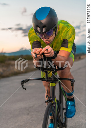 Triathlete riding his bicycle during sunset, preparing for a marathon. The warm colors of the sky provide a beautiful backdrop for his determined and focused effort. Triathlete riding his bicycle during sunset, preparing for a marathon. The warm colors of the sky provide a beautiful backdrop for his determined and focused effort. 115973308