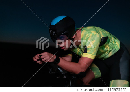 A triathlete rides his bike in the darkness of night, pushing himself to prepare for a marathon. The contrast between the darkness and the light of his bike creates a sense of drama and highlights the A triathlete rides his bike in the darkness of night, pushing himself to prepare for a marathon. The contrast between the darkness and the light of his bike creates a sense of drama and highlights the 115973351