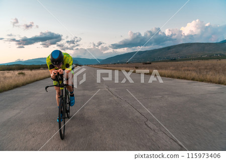 Triathlete riding his bicycle during sunset, preparing for a marathon. The warm colors of the sky provide a beautiful backdrop for his determined and focused effort. Triathlete riding his bicycle during sunset, preparing for a marathon. The warm colors of the sky provide a beautiful backdrop for his determined and focused effort. 115973406
