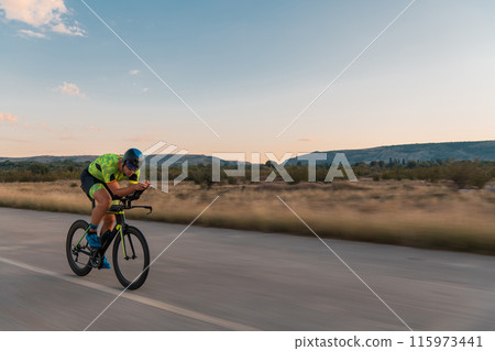 Triathlete riding his bicycle during sunset, preparing for a marathon. The warm colors of the sky provide a beautiful backdrop for his determined and focused effort. 115973441