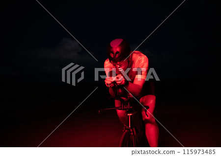 A triathlete rides his bike in the darkness of night, pushing himself to prepare for a marathon. The contrast between the darkness and the light of his bike creates a sense of drama and highlights the 115973485