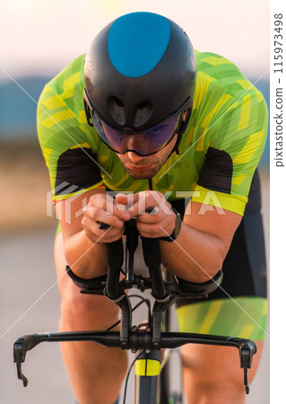 Close up photo of triathlete riding his bicycle during sunset, preparing for a marathon. The warm colors of the sky provide a beautiful backdrop for his determined and focused effort. 115973498