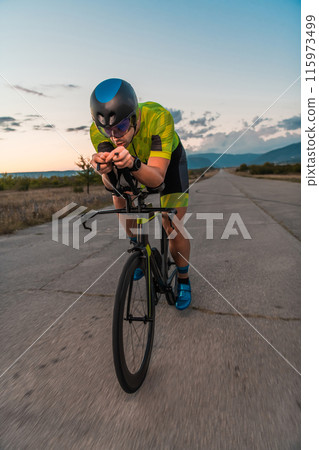 Triathlete riding his bicycle during sunset, preparing for a marathon. The warm colors of the sky provide a beautiful backdrop for his determined and focused effort. Triathlete riding his bicycle during sunset, preparing for a marathon. The warm colors of the sky provide a beautiful backdrop for his determined and focused effort. 115973499