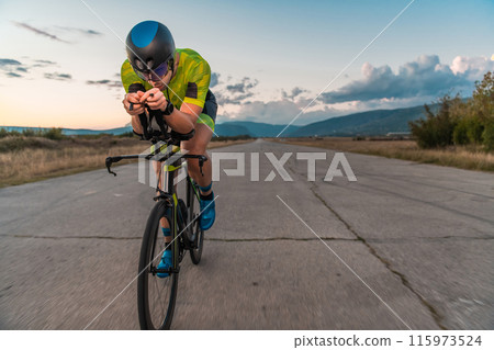Triathlete riding his bicycle during sunset, preparing for a marathon. The warm colors of the sky provide a beautiful backdrop for his determined and focused effort. 115973524