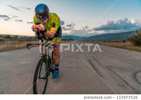Triathlete riding his bicycle during sunset, preparing for a marathon. The warm colors of the sky provide a beautiful backdrop for his determined and focused effort. 115973526