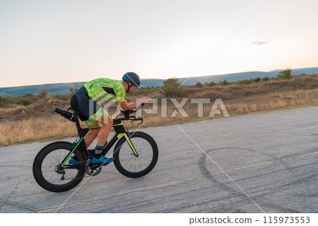 Triathlete riding his bicycle during sunset, preparing for a marathon. The warm colors of the sky provide a beautiful backdrop for his determined and focused effort. 115973553