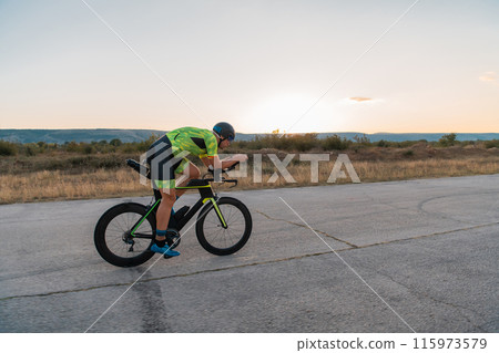 Triathlete riding his bicycle during sunset, preparing for a marathon. The warm colors of the sky provide a beautiful backdrop for his determined and focused effort. Triathlete riding his bicycle during sunset, preparing for a marathon. The warm colors of the sky provide a beautiful backdrop for his determined and focused effort. 115973579