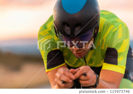 Close up photo of triathlete riding his bicycle during sunset, preparing for a marathon. The warm colors of the sky provide a beautiful backdrop for his determined and focused effort. 115973746