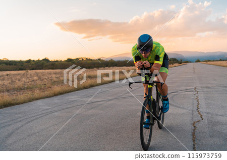 Triathlete riding his bicycle during sunset, preparing for a marathon. The warm colors of the sky provide a beautiful backdrop for his determined and focused effort. Triathlete riding his bicycle during sunset, preparing for a marathon. The warm colors of the sky provide a beautiful backdrop for his determined and focused effort. 115973759