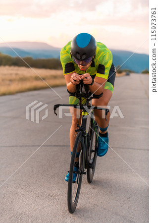Triathlete riding his bicycle during sunset, preparing for a marathon. The warm colors of the sky provide a beautiful backdrop for his determined and focused effort. Triathlete riding his bicycle during sunset, preparing for a marathon. The warm colors of the sky provide a beautiful backdrop for his determined and focused effort. 115973761
