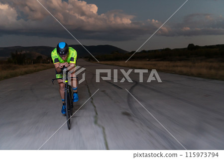 A triathlete rides his bike in the darkness of night, pushing himself to prepare for a marathon. The contrast between the darkness and the light of his bike creates a sense of drama and highlights the A triathlete rides his bike in the darkness of night, pushing himself to prepare for a marathon. The contrast between the darkness and the light of his bike creates a sense of drama and highlights the 115973794