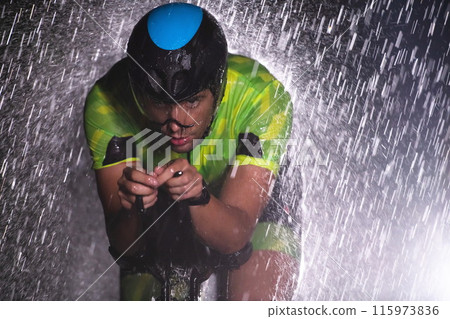 A triathlete braving the rain as he cycles through the night, preparing himself for the upcoming marathon. The blurred raindrops in the foreground and the dark, moody atmosphere in the background add 115973836