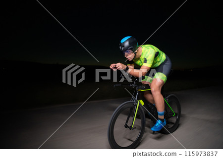 A triathlete rides his bike in the darkness of night, pushing himself to prepare for a marathon. The contrast between the darkness and the light of his bike creates a sense of drama and highlights the A triathlete rides his bike in the darkness of night, pushing himself to prepare for a marathon. The contrast between the darkness and the light of his bike creates a sense of drama and highlights the 115973837