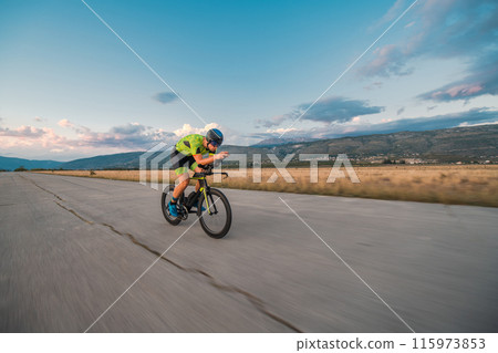 Triathlete riding his bicycle during sunset, preparing for a marathon. The warm colors of the sky provide a beautiful backdrop for his determined and focused effort. 115973853
