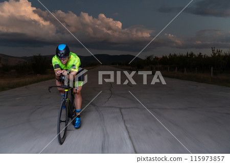 A triathlete rides his bike in the darkness of night, pushing himself to prepare for a marathon. The contrast between the darkness and the light of his bike creates a sense of drama and highlights the A triathlete rides his bike in the darkness of night, pushing himself to prepare for a marathon. The contrast between the darkness and the light of his bike creates a sense of drama and highlights the 115973857