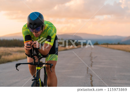 Triathlete riding his bicycle during sunset, preparing for a marathon. The warm colors of the sky provide a beautiful backdrop for his determined and focused effort. 115973869