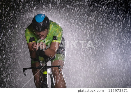 A triathlete braving the rain as he cycles through the night, preparing himself for the upcoming marathon. The blurred raindrops in the foreground and the dark, moody atmosphere in the background add A triathlete braving the rain as he cycles through the night, preparing himself for the upcoming marathon. The blurred raindrops in the foreground and the dark, moody atmosphere in the background add 115973871