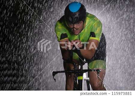 A triathlete braving the rain as he cycles through the night, preparing himself for the upcoming marathon. The blurred raindrops in the foreground and the dark, moody atmosphere in the background add A triathlete braving the rain as he cycles through the night, preparing himself for the upcoming marathon. The blurred raindrops in the foreground and the dark, moody atmosphere in the background add 115973881